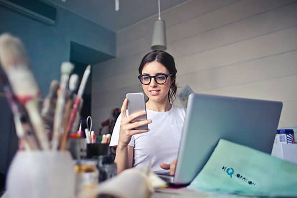 young lady working on laptop looking at phone with art supplies on the desk