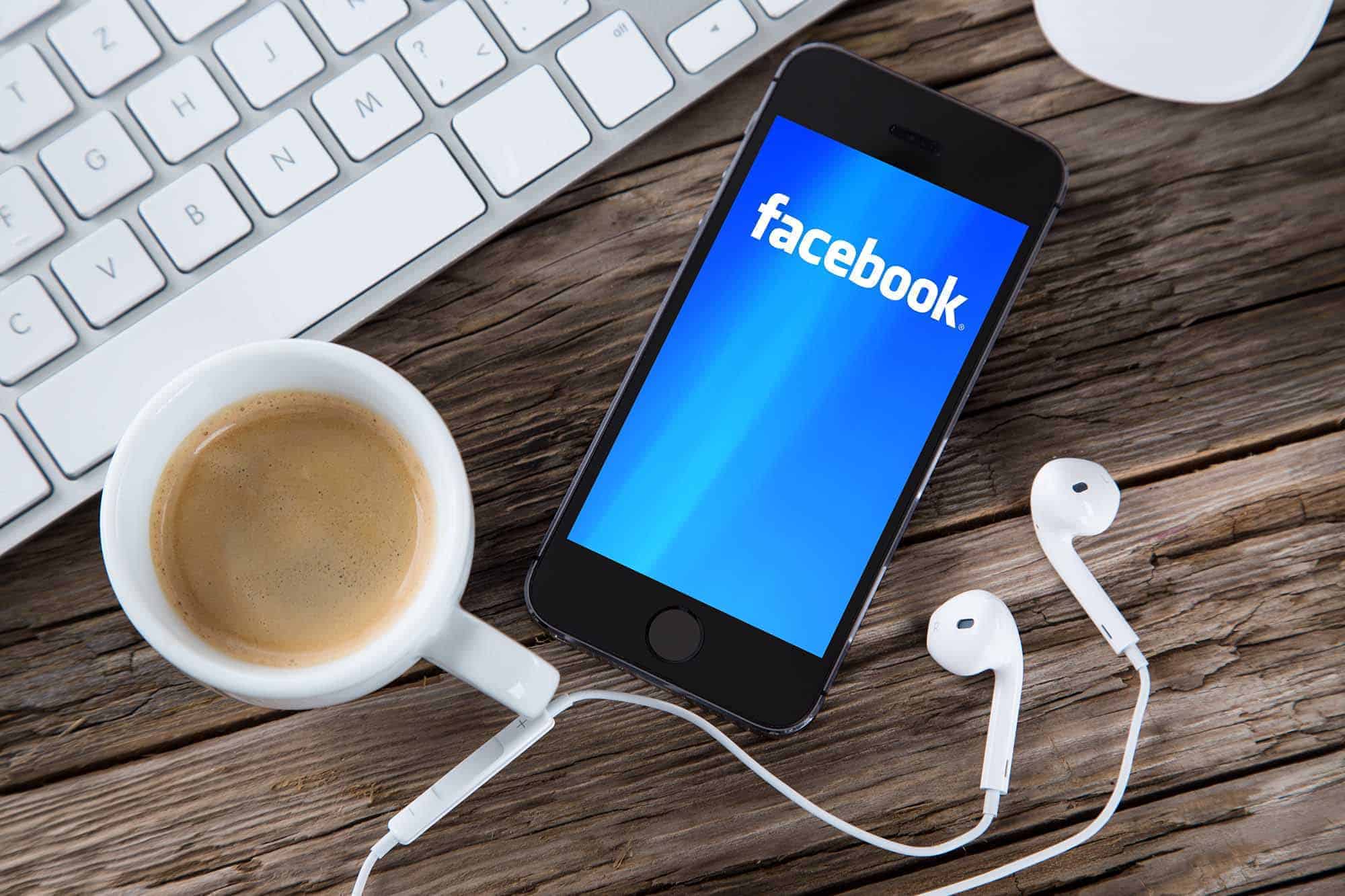Facebook screen on smart phone with headphones, cup of coffee and computer keyboard on desk